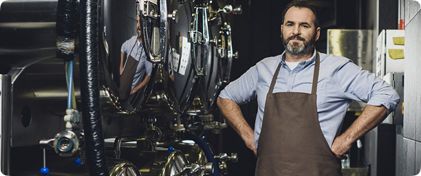 Brewer standing confidently in a brewery, surrounded by stainless steel fermentation tanks, wearing an apron, emphasizing the craft of brewing and the use of CoolBot technology for efficient temperature control.
